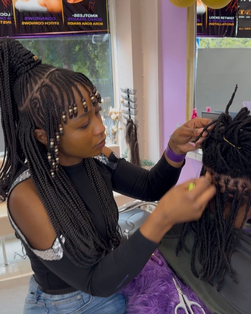 Stylist med Fulani braids och pärlor gör retwist på en kunds locs i salongen, med salongens skyltar i bakgrunden