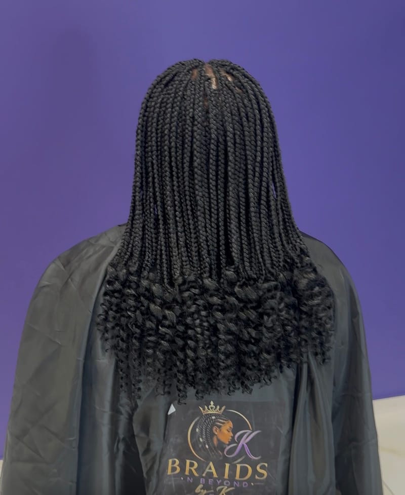 Knotless braids med lockiga toppar, fotograferade bakifrån mot en lila vägg, salongens logotyp på kappan