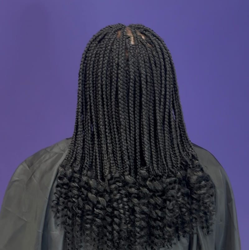 Knotless braids med lockiga toppar mot en lila vägg, salongens logotyp på kappan