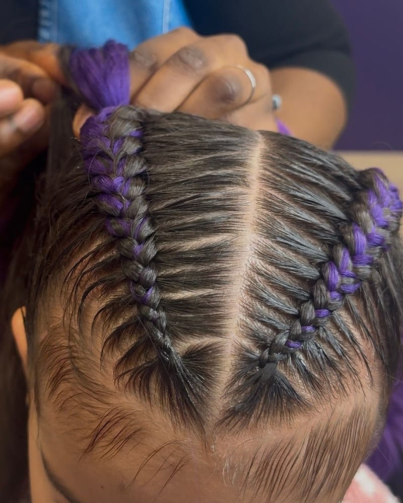 Två cornrows med lila flätinslag, sedda ovanifrån medan stylisten flätar